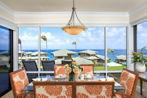 Un coin repas avec une table dressée pour quatre personnes, de grandes fenêtres offrant une vue sur l’océan et les bâtiments du complexe hôtelier, et un luminaire suspendu au-dessus.