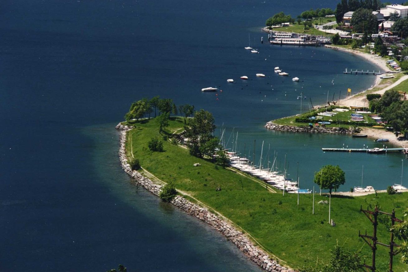 Vue aérienne d’un port de plaisance situé sur la côte, avec des bateaux amarrés le long du rivage, entouré de verdure et de bâtiments résidentiels.