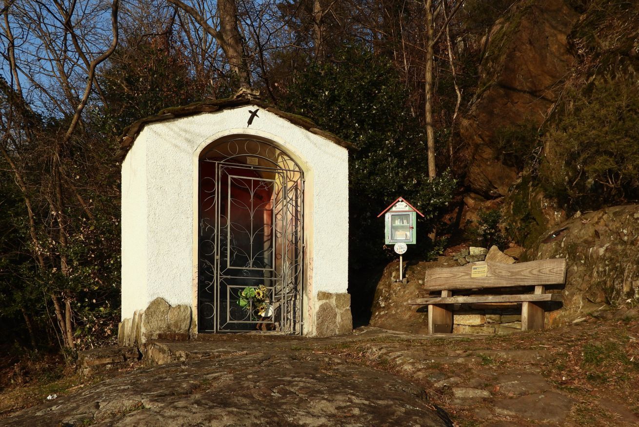 Une petite chapelle blanche, dotée d’une porte en métal et surmontée d’une croix, entourée d’arbres et bordée d’un banc en pierre.
