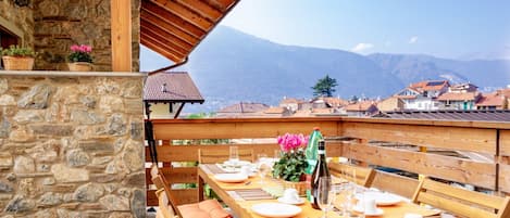 A wooden dining table set for four with a stone wall backdrop, a bottle, and a view of a mountain range.