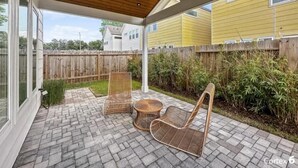 Eine überdachte Terrasse mit Korbmöbeln und einem Ziegelboden, mit Blick auf einen Garten mit Holzzaun und Bepflanzung.
