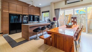 A modern kitchen with a central island, bar stools, and a dining area with a wooden table and chairs. - Modern 10BR ZipLine 3 Pools  V256 (Rawai)