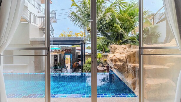 A view through a glass door of a swimming pool area with a tiled floor, a rock formation, and a dining area with tables and chairs. - Modern 10BR ZipLine 3 Pools  V256 (Rawai)