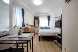 Ein kleines Zimmer mit Bett, Schreibtisch, Stuhl, einem kleinen Tisch mit Pflanze, einer Mikrowelle und einem Regal mit Geschirr.