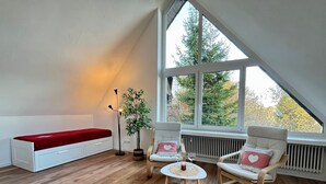 Un acogedor dormitorio tipo loft con techo inclinado, una cama roja, dos mecedoras, una mesita con una vela y un ventanal con vistas a árboles.