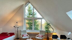 Un dormitorio tipo loft acogedor con un tragaluz grande, una cama con ropa de cama roja, dos mecedoras, una mesita redonda con bebidas y una mesa de comedor con tazas y platillos.