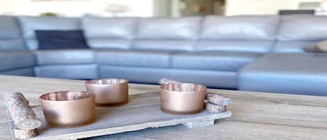 A wooden tray with three copper-colored cups and a decorative item on a table in a living room with a grey sofa.