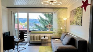 Ein modernes Wohnzimmer mit einer Couch, einem Couchtisch und einem Fernseher. Ein Fenster bietet Blick auf Bäume und Gebäude.