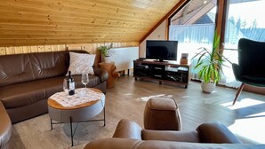 Ein gemütliches Wohnzimmer mit einem braunen Ledersofa, einem kleinen runden Couchtisch, einem Fernseher auf einem Holzboden und einem großen Fenster mit Blick auf Bäume.