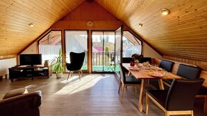 Ein gemütliches Dachzimmer mit schrägverkleidetem Holzboden, einem Esstisch für vier Personen, einem Fernseher und einem Topfpflanzen.