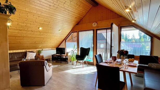 Ein geräumiges Loft mit hohen Decken, Holzbalken, einer gemütlichen Sitzecke mit Sofa und Sesseln, einem Esstisch für zwei Personen und großen Fenstern mit Blick ins Freie.