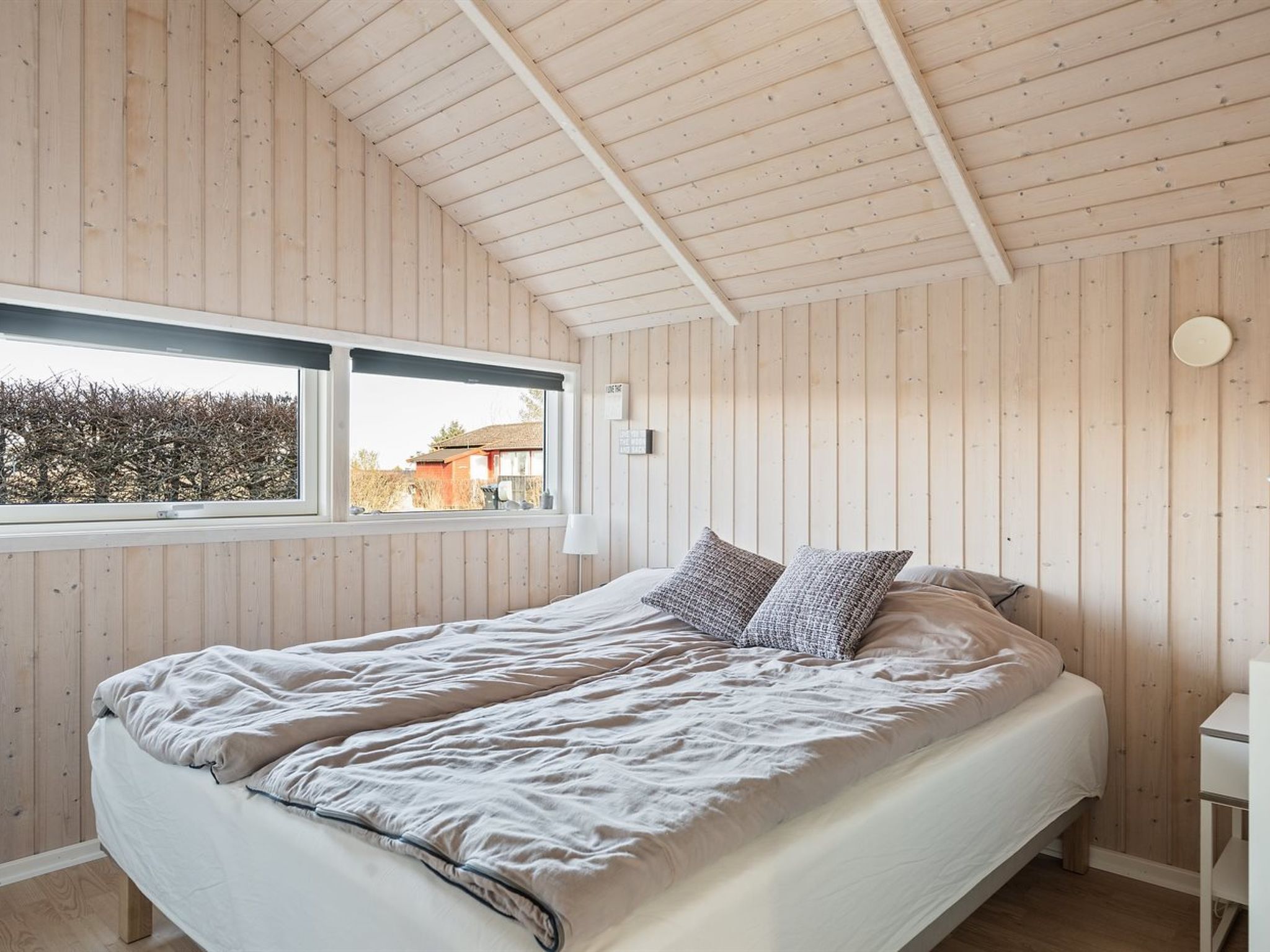 Une chambre avec un plafond en bois à lattes, un grand lit avec un matelas blanc et deux coussins à motifs, une fenêtre donnant sur une maison et une table de chevet blanche.