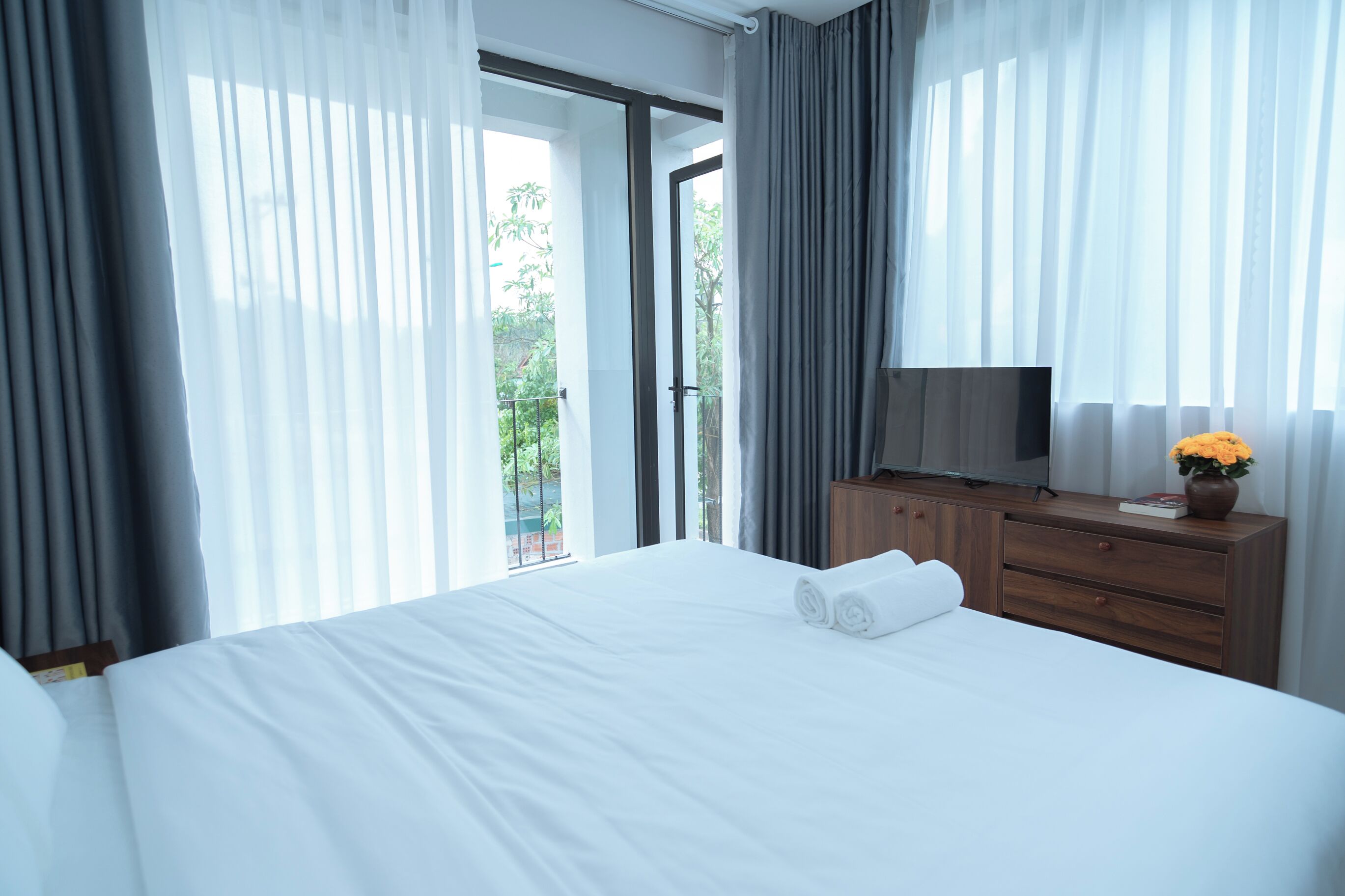A hotel room with a large bed, a wooden dresser, a flat-screen TV, and a view of a balcony with greenery.