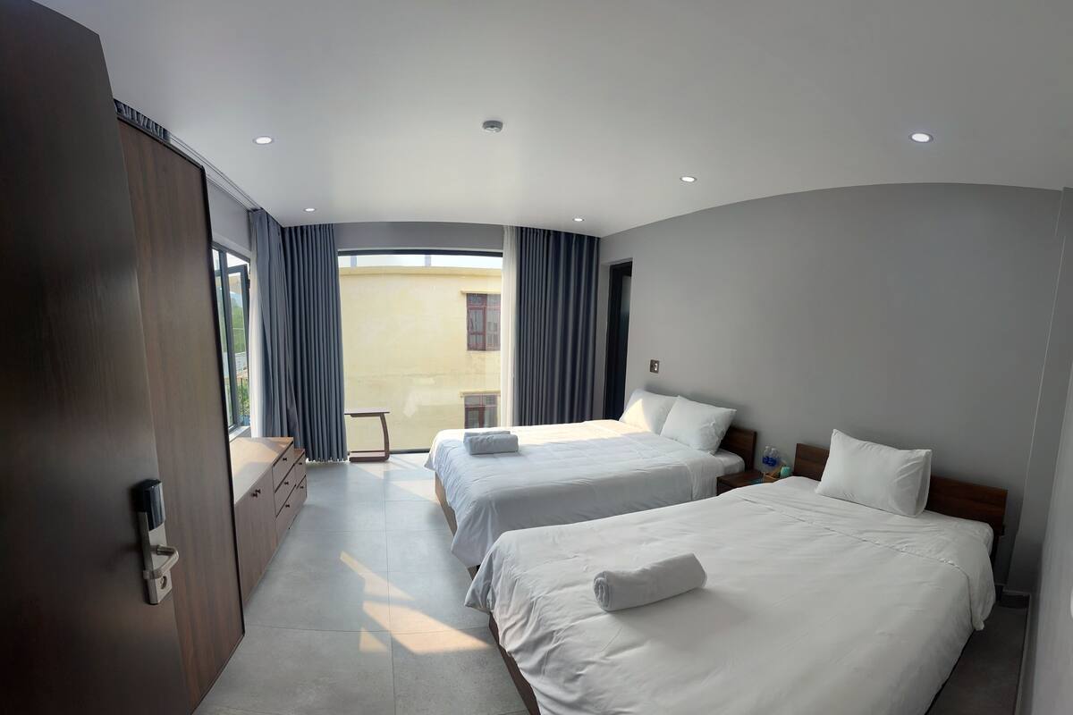 Ein Hotelzimmer mit zwei Betten, einem Schreibtisch und Blick auf ein Gebäude durch das Fenster.