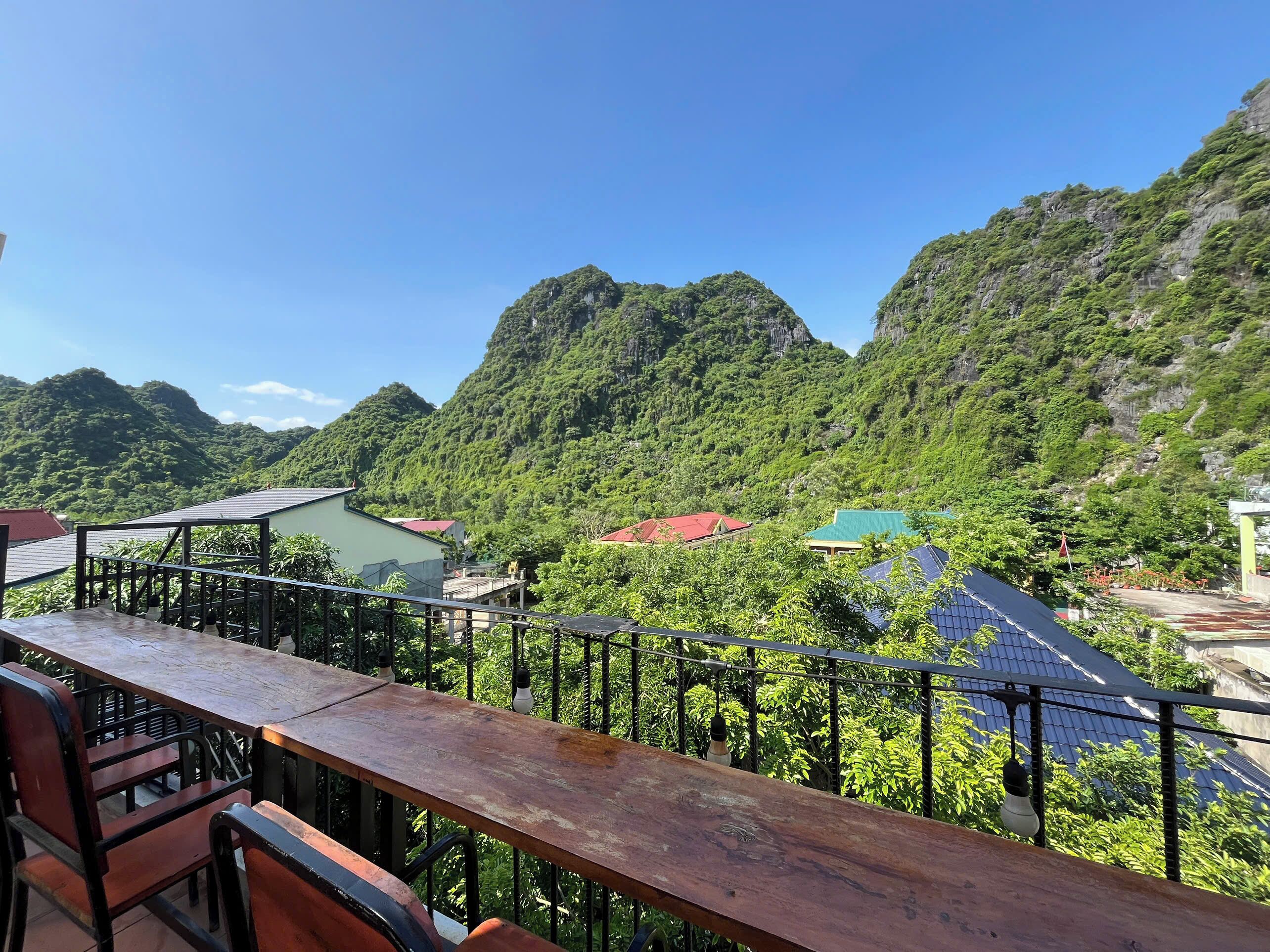 A terrace with a wooden table and chairs, overlooking a mountainous landscape with lush greenery and buildings with red and green roofs.