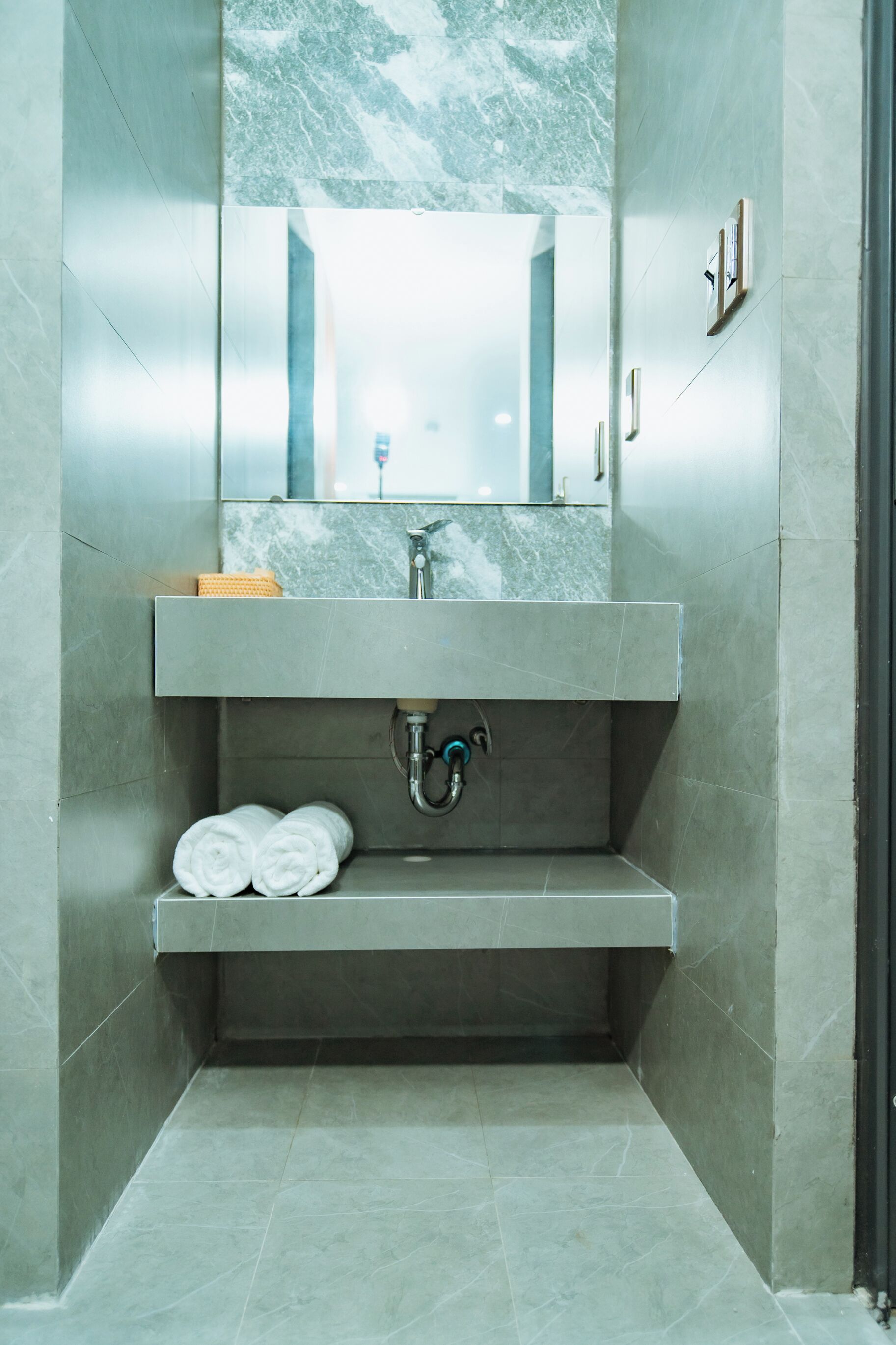 A modern bathroom with a marble sink, a mirror, and two rolled towels on a shelf.