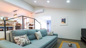 Una sala de estar con sofá, mesa de centro y televisor. Se ve una escalera que sube a un nivel superior con cocina y comedor.