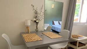 A modern dining area with a table, chairs, a vase with branches, a lamp, and a mirror reflecting a bed. - Detached studio with terrace in Aix countryside property (Aix-en-Provence)