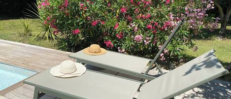 Une chaise longue installée au bord de la piscine, avec un chapeau et des lunettes de soleil posés dessus, une piscine et, en arrière-plan, des fleurs roses en pleine floraison.