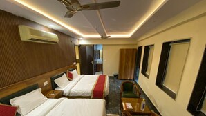 A hotel room with two beds, a ceiling fan, an air conditioning unit, a small table with a chair, and a view of the hallway. - Hotel Home Stay Residency (New Delhi)