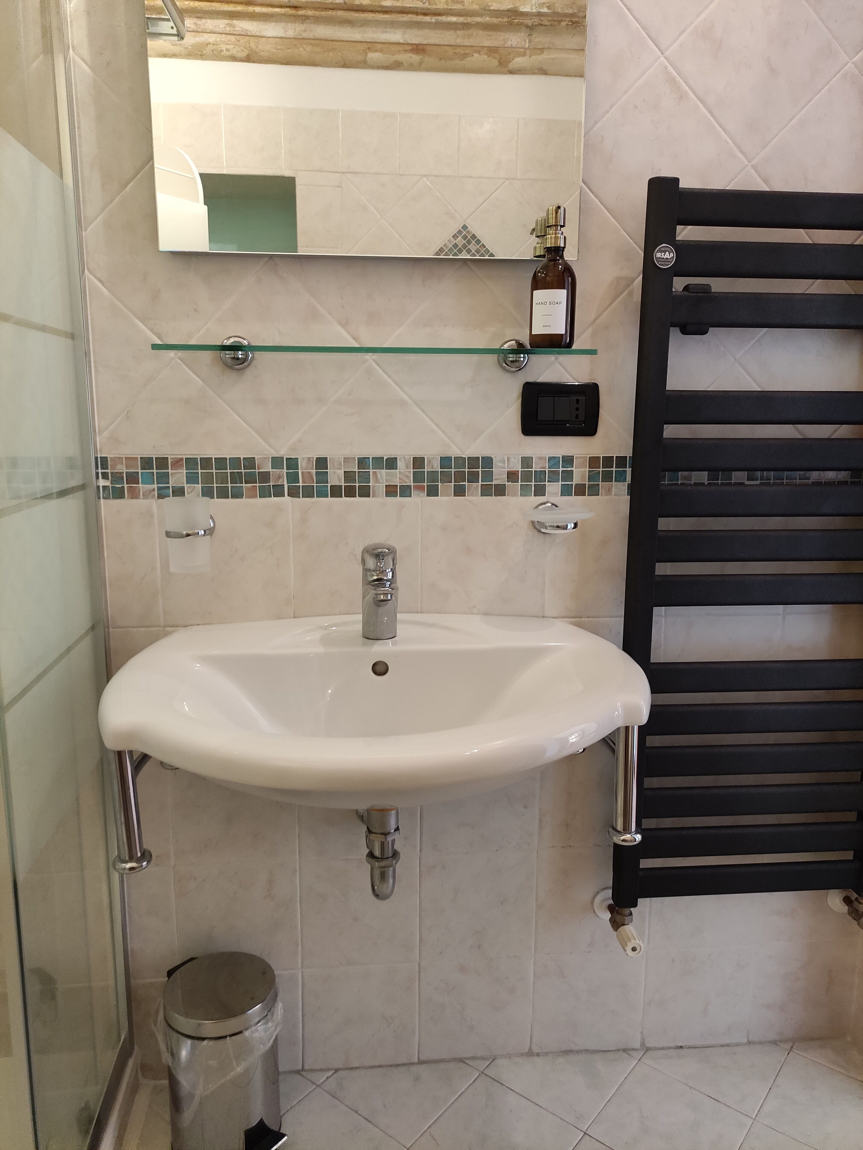 Un baño con lavamanos blanco, repisa de vidrio encima, toallero negro y salpicadero de azulejos.