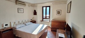 Ein Schlafzimmer mit Bett, Nachttisch, Lampe, Kommode, Fenster und einem an der Wand hängenden Kunstwerk.