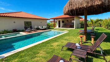 Une piscine avec des chaises longues, un parasol en chaume et un espace patio comprenant une table et des chaises.