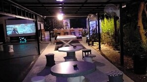 Un espace extérieur aménagé avec des tables et des tabourets, un grand écran diffusant un film, et un bar avec un comptoir et des tabourets.