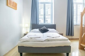 Ein ordentlich bezogenes Bett mit weißen Leinen und einem Kissen, eine Nachttischlampe und Blick auf Gebäude durch ein Fenster mit blauen Vorhängen.