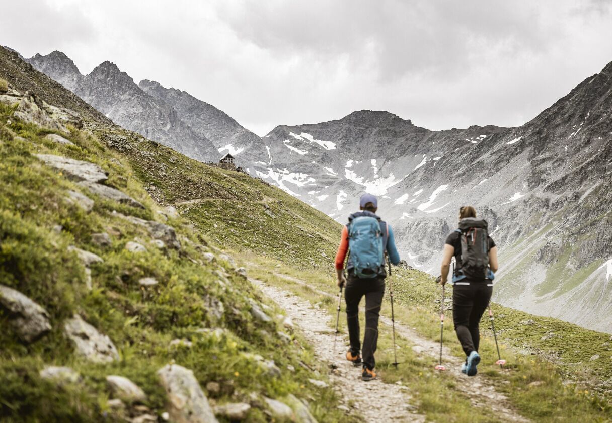Zwei Wanderer auf einem Bergpfad mit felsigem Gelände und spärlicher Vegetation, umgeben von steilen, schneebedeckten Bergen.
