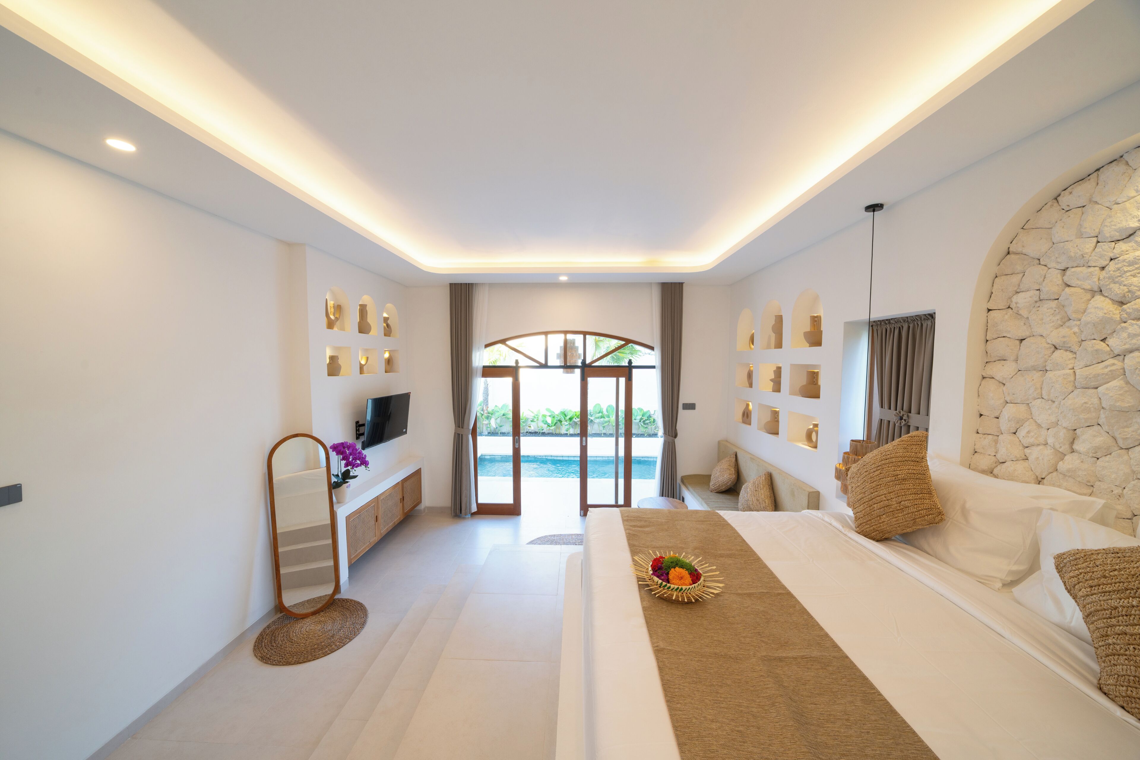 A modern hotel room with a large bed, a flat-screen TV, a vanity with a mirror, and a stone accent wall.
