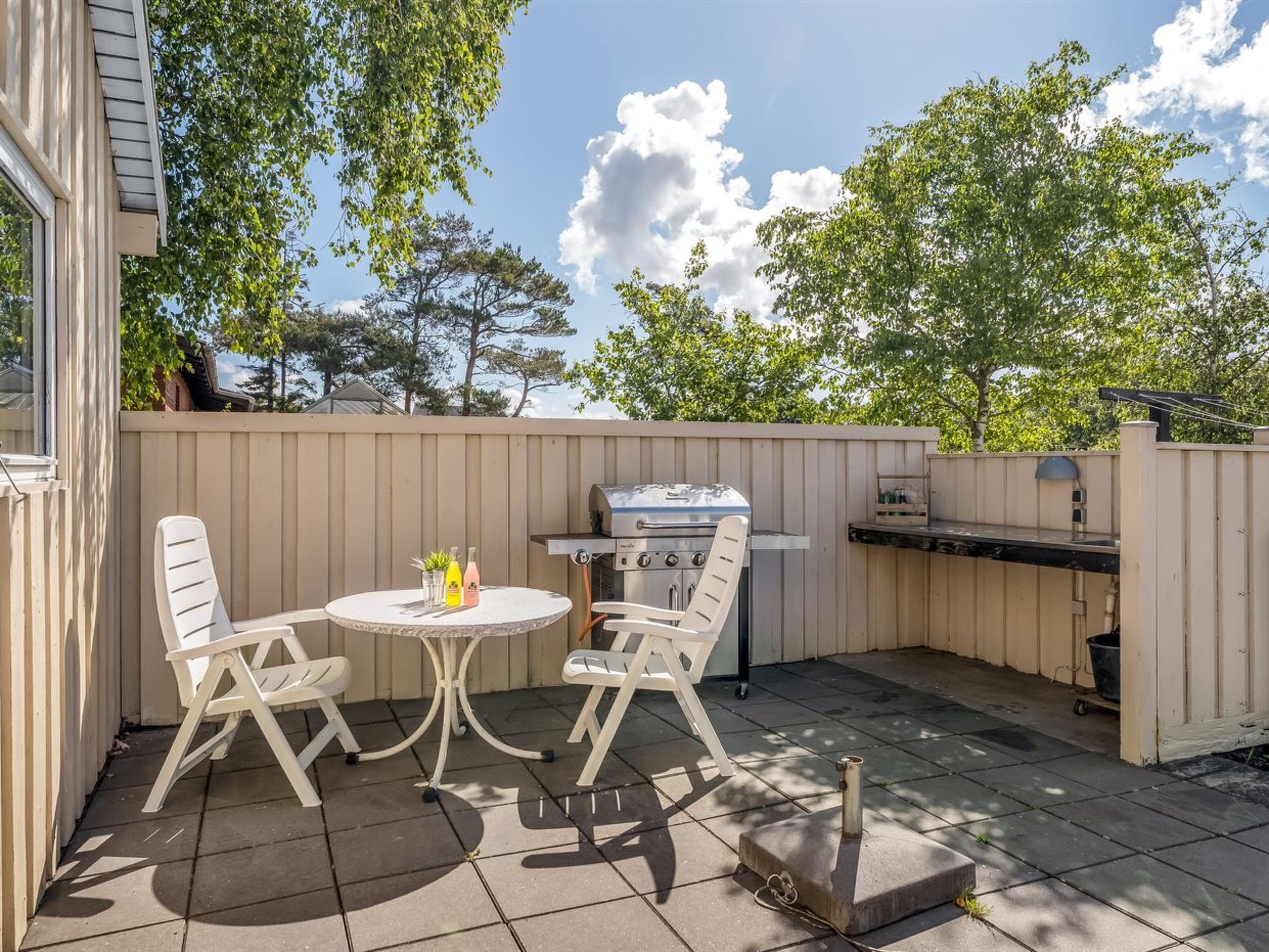 Een terras buiten met een ronde tafel en twee stoelen, een grill en een klein barbecuegedeelte.