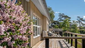 Una terraza de madera con mobiliario exterior, baranda y una casa con revestimiento vertical y grandes ventanales.