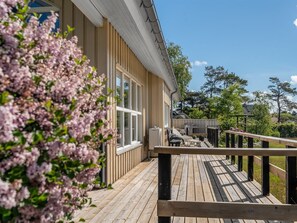 Una terraza de madera con mobiliario exterior, baranda y una casa con revestimiento vertical y grandes ventanales.