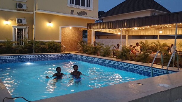 A swimming pool with two people in the water, surrounded by a covered patio area with string lights. - Rossy's Apartments Serviced Apts Abuja (Abuja)