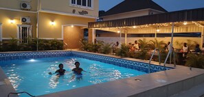 A swimming pool with two people in the water, surrounded by a covered patio area with string lights. - Rossy's Apartments Serviced Apts Abuja (Abuja)
