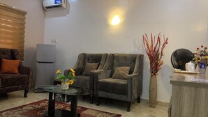 A living room with a sofa, a coffee table with flowers, a fan, and a refrigerator. - Rossy's Apartments Serviced Apts Abuja (Abuja)