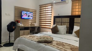A hotel room with a bed, a desk, a chair, a TV, and a standing fan. - Rossy's Apartments Serviced Apts Abuja (Abuja)