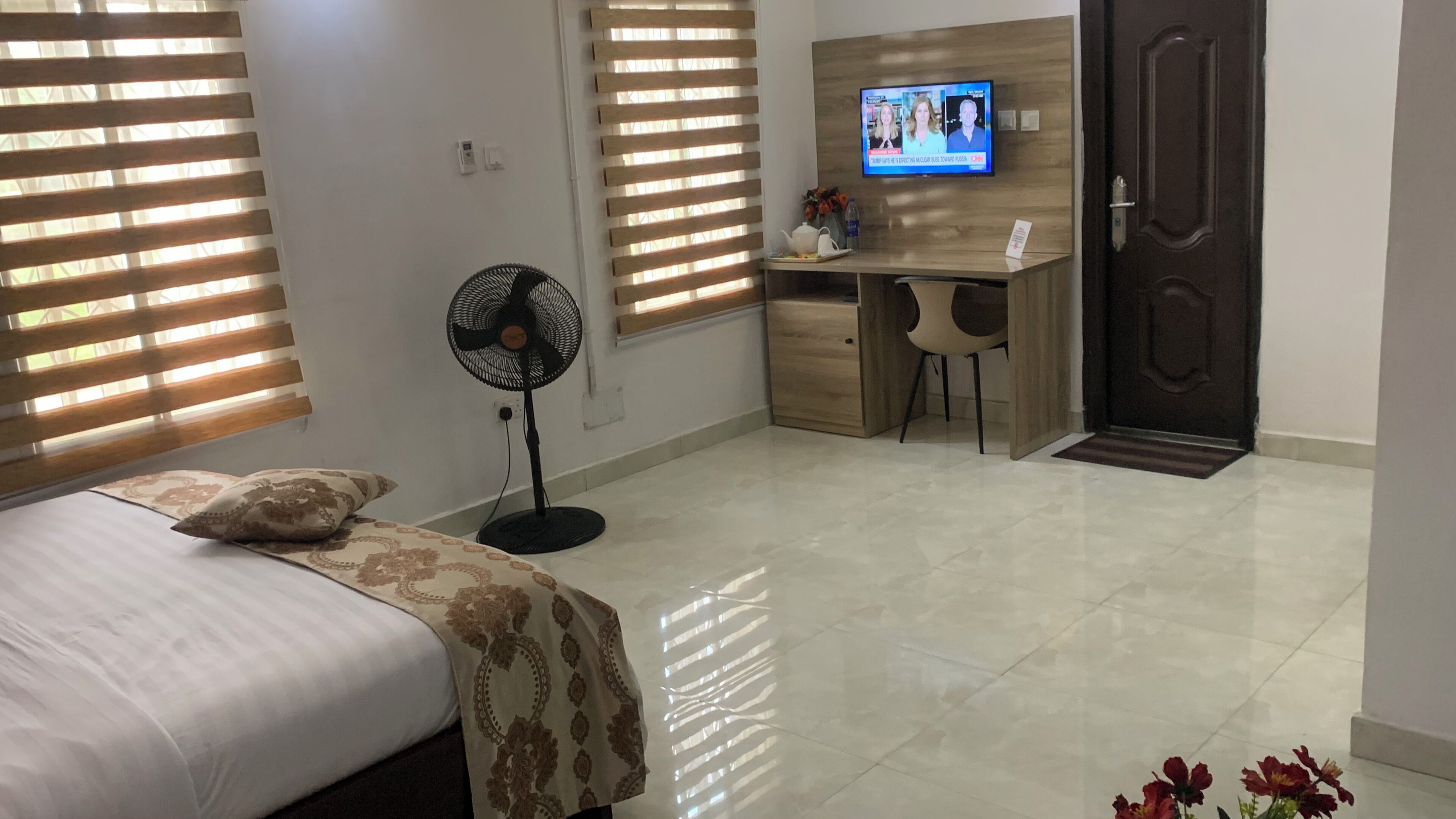 A hotel room with a bed, a desk, a chair, a TV, and a fan.