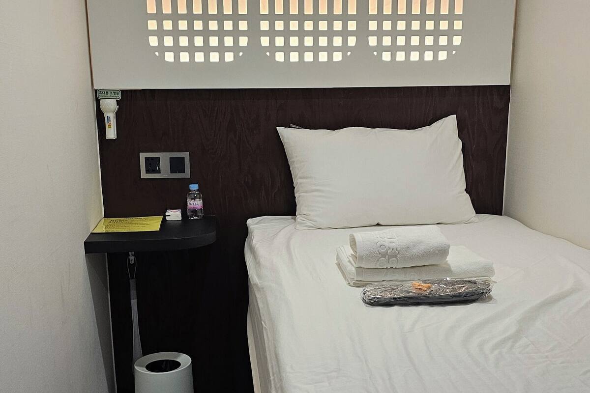 Una habitación de hotel con una cama bien hecha, una mesita de noche con una botella y una caja de pañuelos, y una lámpara fijada en la pared.