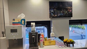 Een hotel receptie met een koffieapparaat, een waterdispenser, een zakdoekendoos, een fles handdesinfectiemiddel en een raam met uitzicht op de stad.