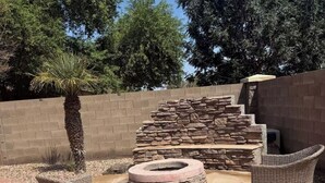 An outdoor patio area with stone flooring, a stone fire pit, wicker furniture, and a palm tree. - Cozy 4bd3ba Pool/FirePit/BBQ House in Maricopa (Maricopa)