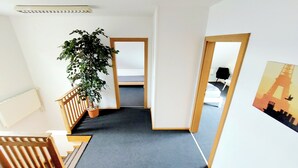 A hallway with a dark carpet, a wooden railing, a potted plant, and a bed in the background. - Guesthouse Graz Central for 9 People (Graz)