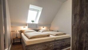 Une chambre avec un plafond en pente, un puits de lumière, un lit avec une literie blanche, une table de chevet avec une lampe et une petite plante.