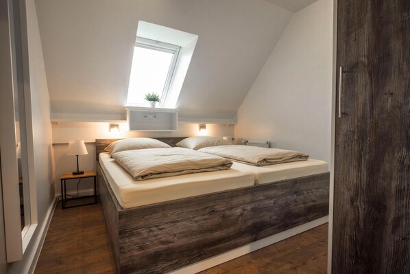 A bedroom with a sloped ceiling, a skylight, a bed with white bedding, a bedside table with a lamp, and a small plant. - Cozy vacation apartment Deichkoje 3 in Dagebüll with modern furnishings (Dagebüll)