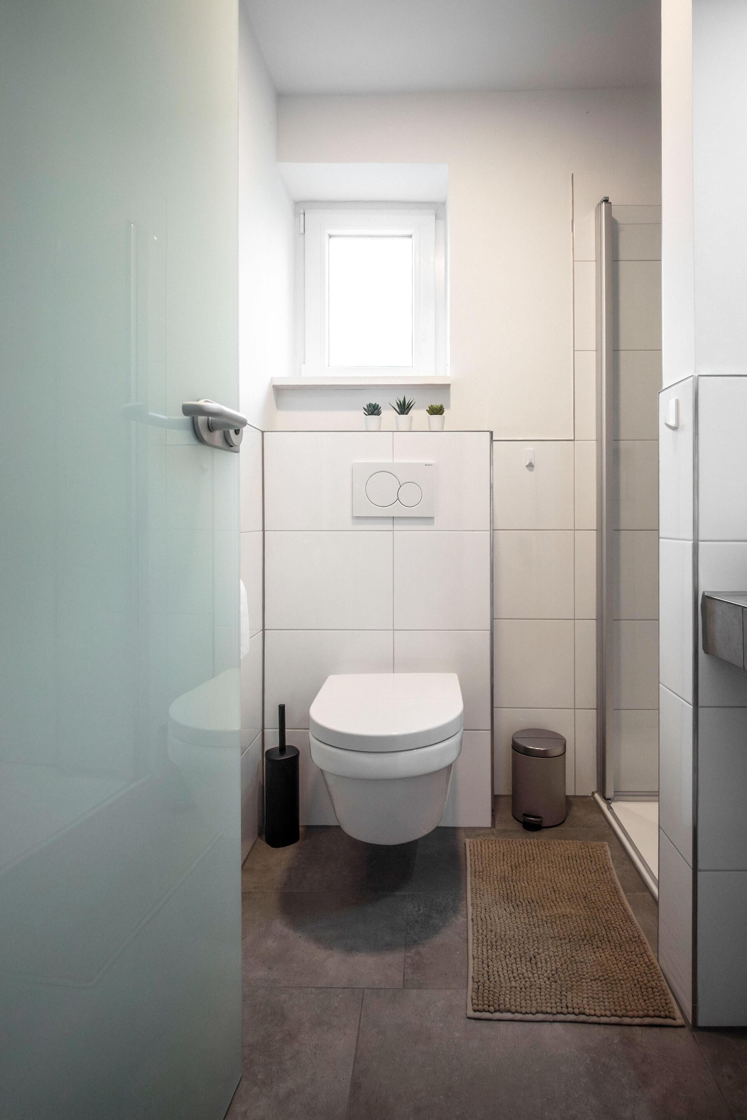 Een compacte badkamer met toilet, een klein raam boven de wastafel en een douchegedeelte.