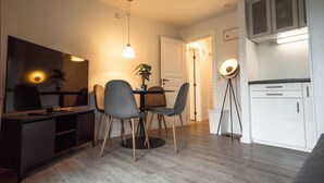 Ein modernes Wohnzimmer mit Flachbildfernseher, einem Esstisch mit Stühlen, einer Stehlampe und einer Küchenzeile mit Schränken.