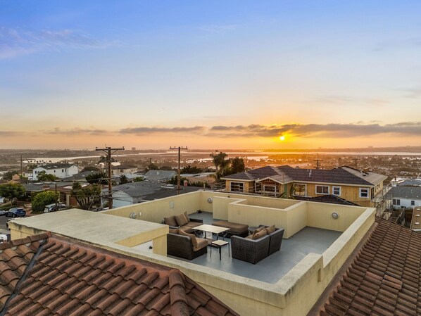 A rooftop terrace with outdoor furniture, a view of the sunset, and surrounding residential buildings. - Tuscan Sands by Coastline Vacation Rentals (San Diego)