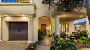 A residential building with a dark garage door, a covered entrance with columns, and a well-maintained garden with various plants and a pathway. - Tuscan Sands by Coastline Vacation Rentals (San Diego)