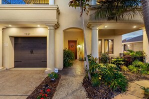 A residential building with a dark garage door, a covered entrance with columns, and a well-maintained garden with various plants and a pathway. - Tuscan Sands by Coastline Vacation Rentals (San Diego)